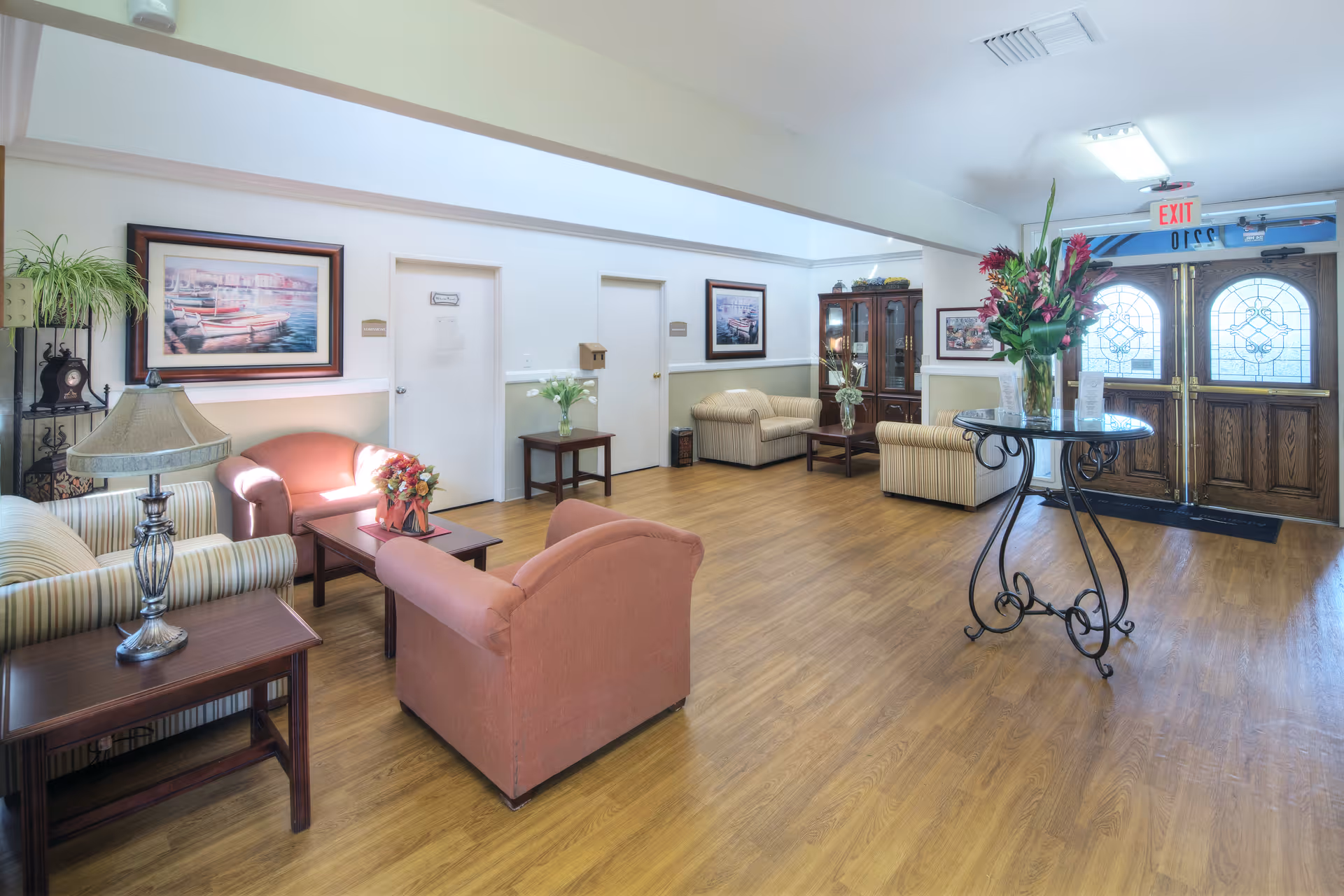 A bright and spacious living room area with wooden flooring, multiple armchairs and sofas arranged around coffee tables, a decorative table with a vase of flowers near the entrance, framed artwork on the walls, and large double doors with stained glass panels letting in natural light.