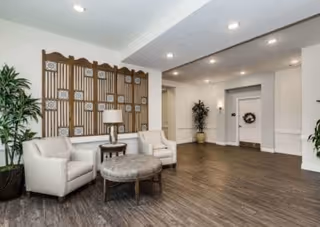 A spacious interior common area with two white armchairs, a round ottoman, a small side table with a lamp, decorative wall art, and potted plants. The floor is wood, and the walls are painted white with recessed lighting in the ceiling.