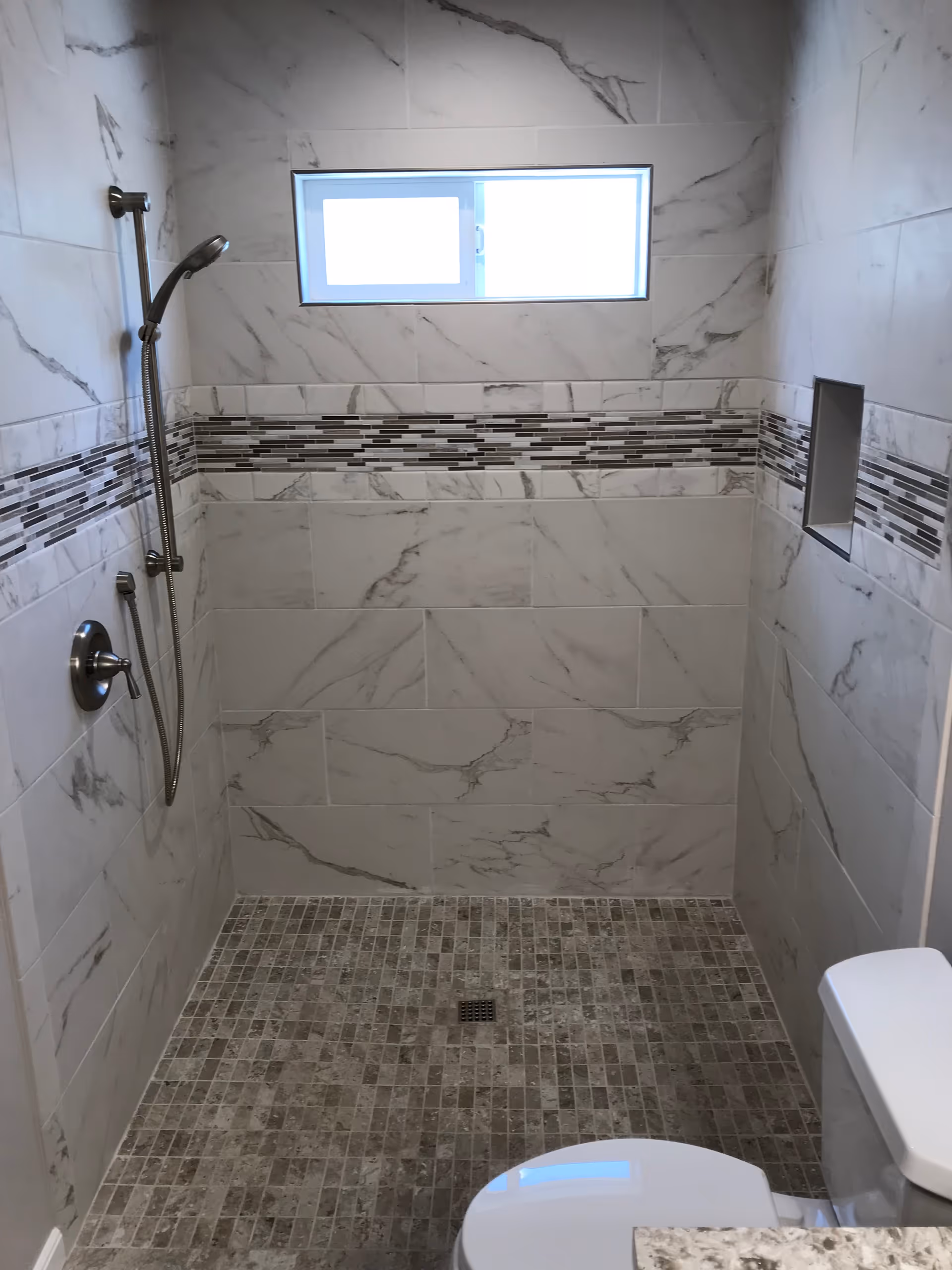 A modern bathroom shower area with marble-patterned wall tiles and a horizontal decorative strip of small rectangular tiles. There is a handheld showerhead mounted on the left wall, a small recessed shelf on the right wall, and a small window near the top center of the shower. The floor is covered with small square tiles in neutral tones. A white toilet is partially visible in the bottom right corner.