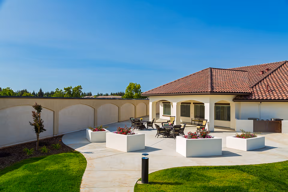 Outdoor patio area at The Gardens At Laguna Springs featuring several black metal tables and chairs with cushions, surrounded by white rectangular planters filled with colorful flowers. The patio is adjacent to a building with a red tile roof and beige walls, under a clear blue sky.