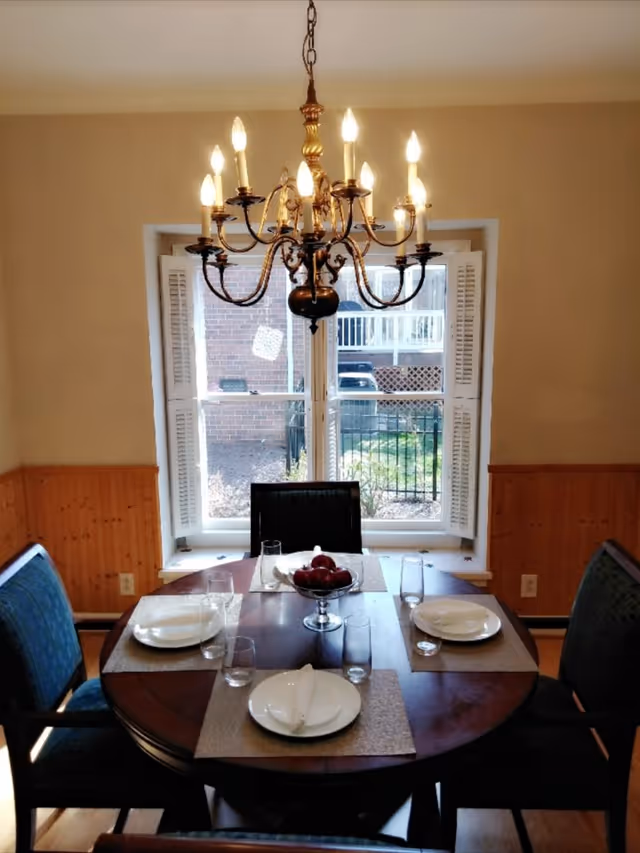 A dining room with a round wooden table set for four people with white plates, glasses, and napkins. A decorative bowl with red apples is in the center of the table. Above the table hangs an ornate chandelier with lit bulbs. Behind the table is a window with white shutters, showing an outdoor view of a brick wall, a fence, and some greenery.
