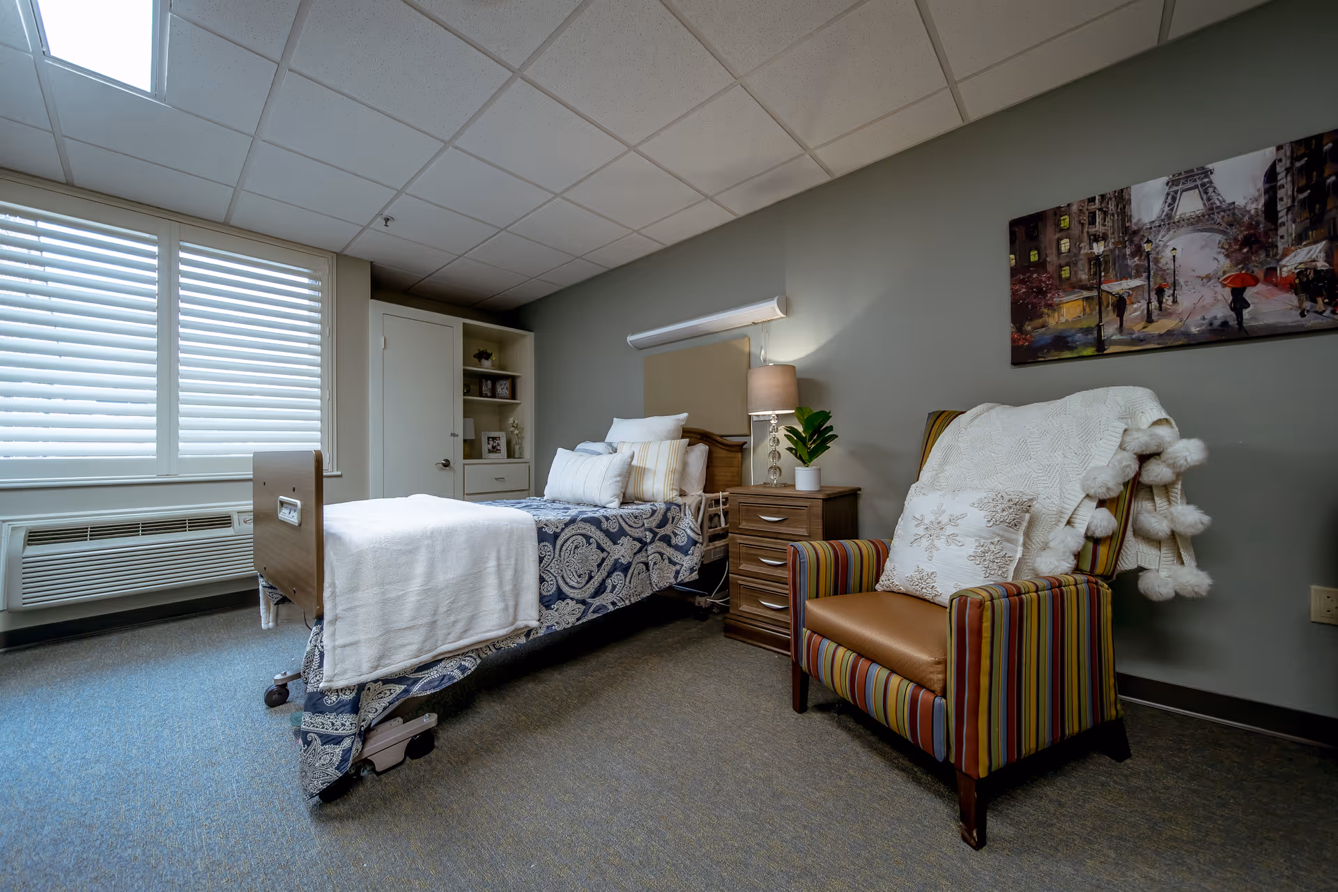A nursing facility bedroom with a single bed covered in a blue and white patterned blanket and white pillows. Next to the bed is a wooden nightstand with a lamp and a small potted plant. A striped armchair with a white throw blanket and decorative pillow is positioned nearby. The room has a window with white blinds, a built-in cabinet, and a painting of a city street scene with the Eiffel Tower on the wall.