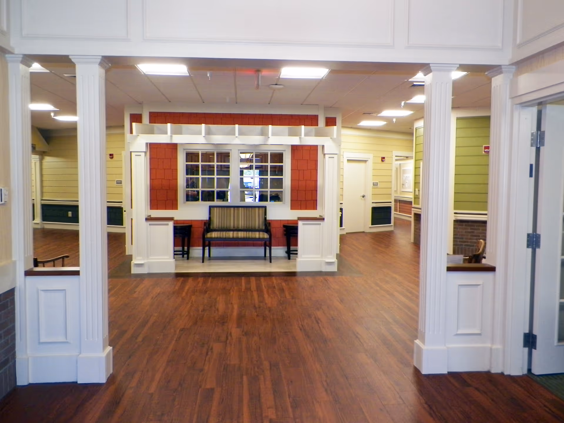 Open interior lobby with hardwood floors, white columns, and a bench seating area against a decorative window wall.