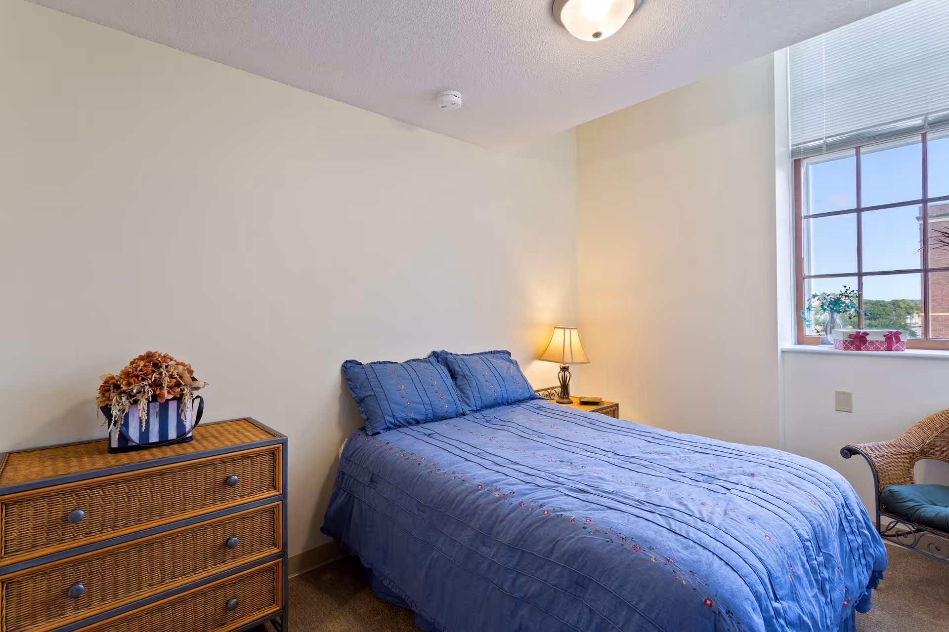 A bedroom with a bed covered in a blue quilt and matching pillows. There is a wicker dresser with a decorative flower arrangement on top to the left of the bed. A small bedside table with a lamp is on the right side of the bed. A window with blinds lets in natural light, and a wicker chair with a cushion is positioned near the window.