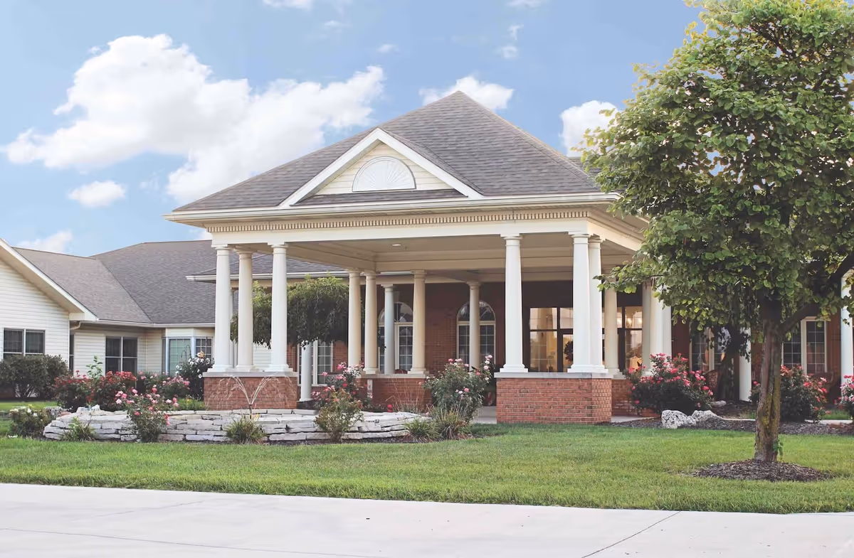 Front exterior view of Cedarhurst Senior Living of Granite City showing a covered entrance with white columns, brick base, manicured lawn, flower beds with pink roses, and a tree on the right side under a partly cloudy sky.