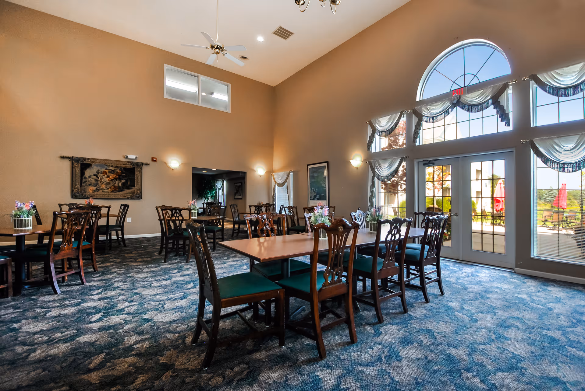 A spacious dining room with multiple wooden tables and chairs arranged neatly. The room features high ceilings with a ceiling fan, large arched windows with sheer curtains allowing natural light to fill the space, and a carpeted floor with a blue and gray pattern. There are small flower arrangements on the tables and artwork on the walls. Outside the windows, patio furniture and red umbrellas are visible.
