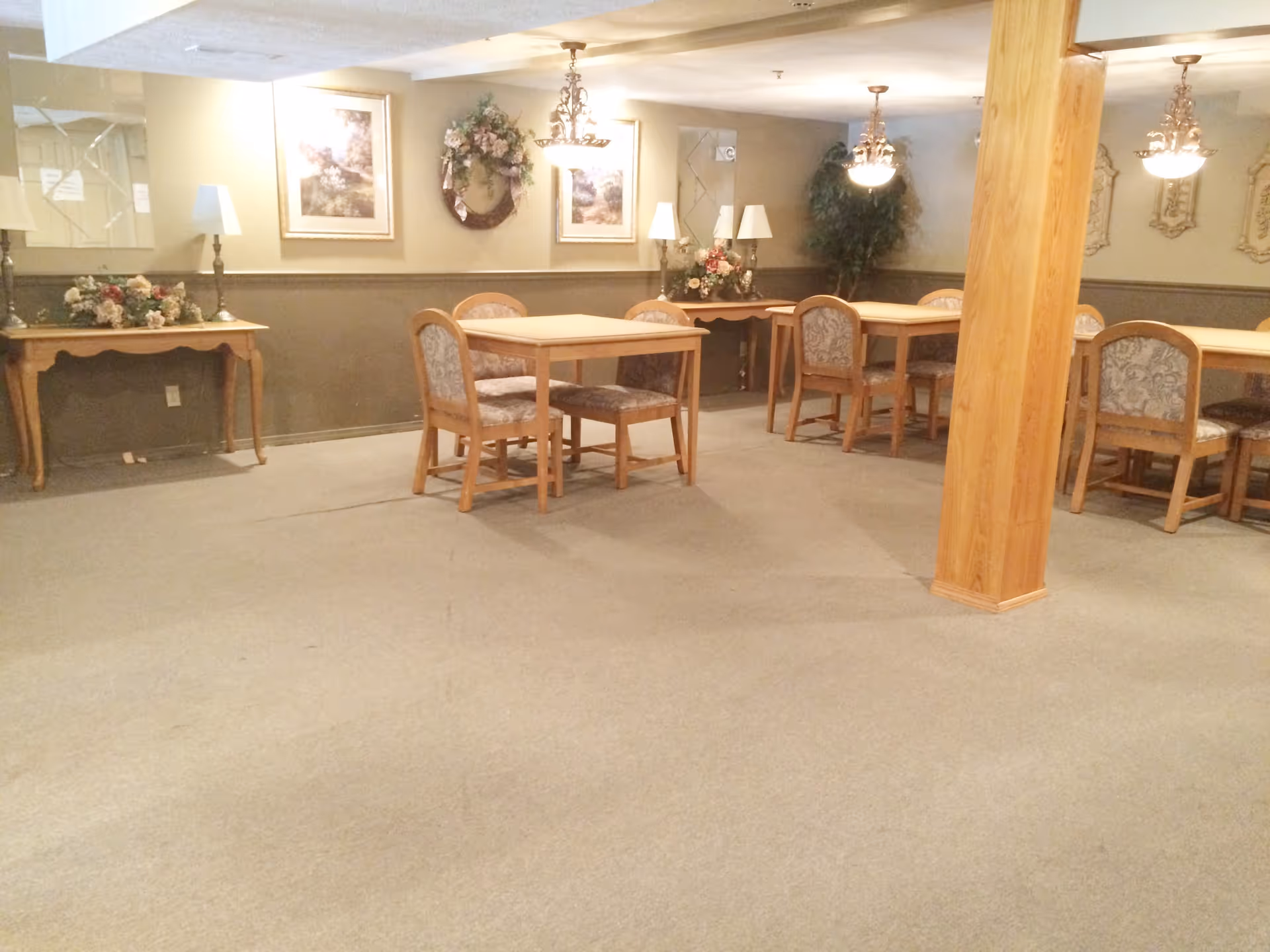 Interior view of a dining area in a senior living facility with several wooden tables and upholstered chairs arranged on a carpeted floor. The walls are decorated with framed pictures, a floral wreath, and floral arrangements on side tables. The room is softly lit by ceiling lights and table lamps, with a wooden pillar visible in the foreground.