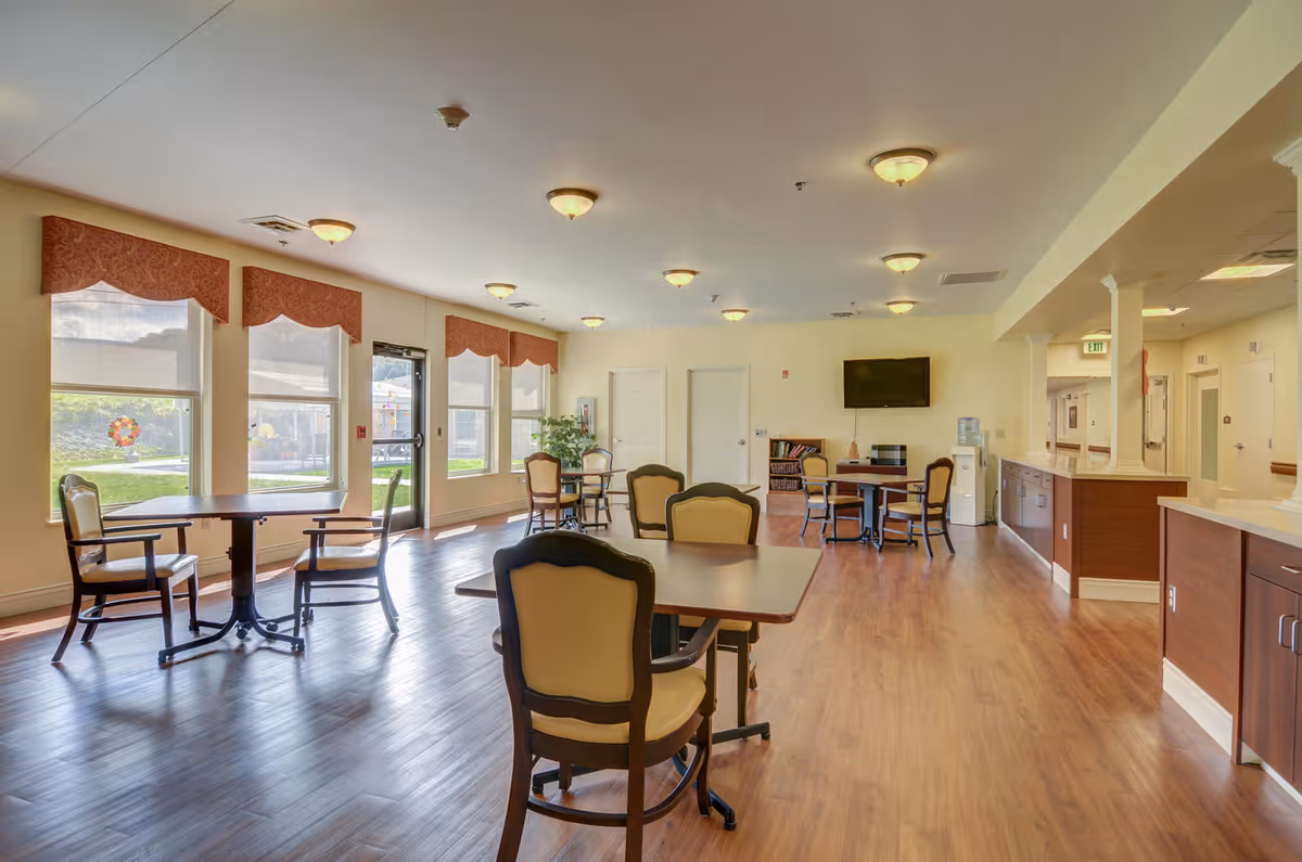 A spacious and well-lit common area in a senior living facility with several tables and chairs arranged for seating. Large windows with red valances allow natural light to fill the room. The floor is wooden, and the walls are painted a light color. There is a flat-screen TV mounted on the far wall, a water dispenser, and a small bookshelf. The room has ceiling lights and a door leading outside.