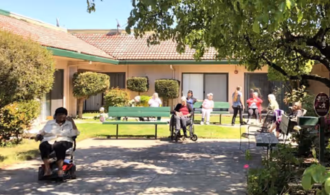 Outdoor courtyard area at Legacy Oaks of Sacramento with several elderly residents sitting on benches and in wheelchairs, enjoying the sunny day. The courtyard is surrounded by a single-story building with a tiled roof and trimmed bushes. Trees provide shade over the paved walkway.