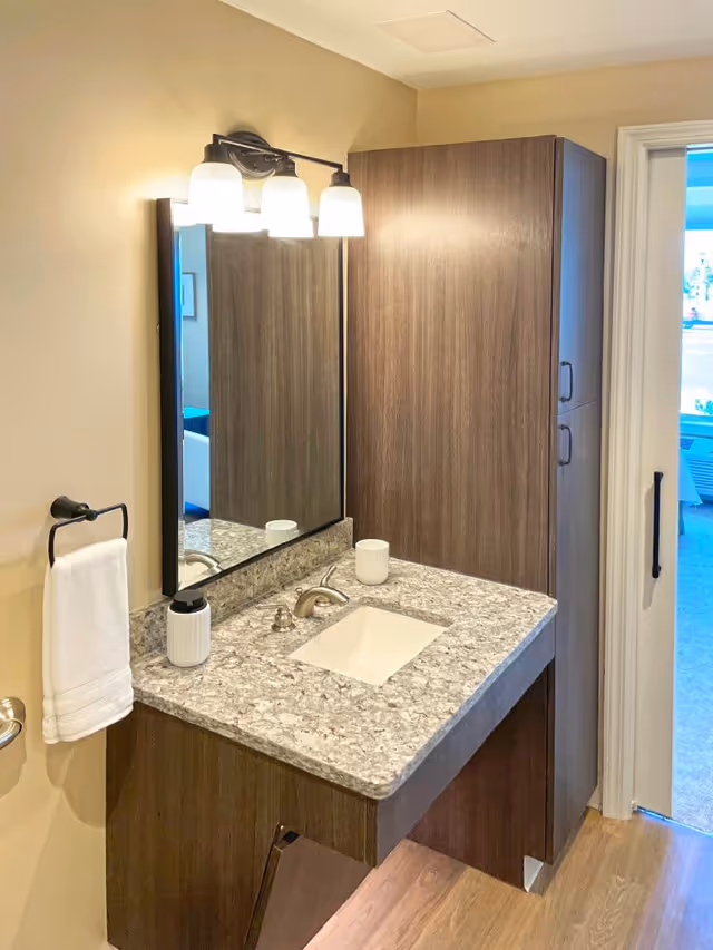 A bathroom vanity with a granite countertop, an under-mount sink, a modern faucet, a soap dispenser, and a cup. Above the sink is a rectangular mirror with three light fixtures mounted on the wall. To the right of the vanity is a tall wooden cabinet. A white towel hangs on a black towel ring on the left wall. The bathroom door is open, showing a glimpse of an adjacent room with a window and outdoor view.