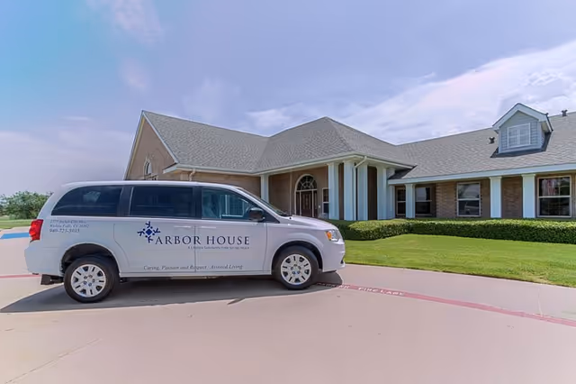 A white van with the logo and text 'Arbor House' parked in front of a single-story brick building with a gray roof and white columns. The building is surrounded by green grass and a clear blue sky is visible.