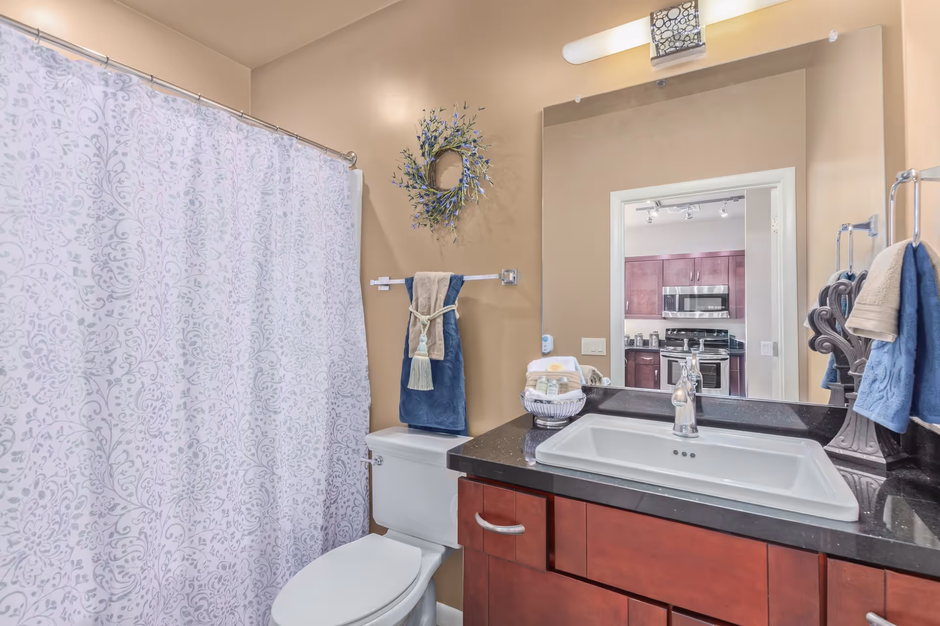 A bathroom with beige walls featuring a white toilet and a shower with a white patterned curtain. There is a dark countertop with a white sink and a silver faucet. Above the sink is a large mirror reflecting a kitchen area with wooden cabinets and stainless steel appliances. Towels hang on the wall, and a decorative wreath is mounted above the toilet.
