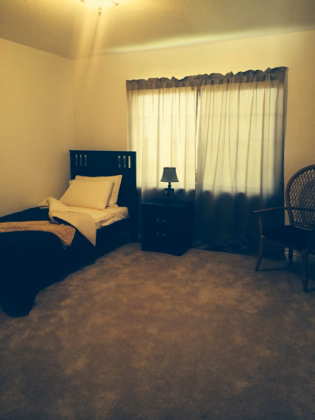 A simple bedroom with a single bed made with white and dark bedding, a dark wooden nightstand with a small lamp, a window covered with beige curtains, and a wicker chair with a cushion on the right side.
