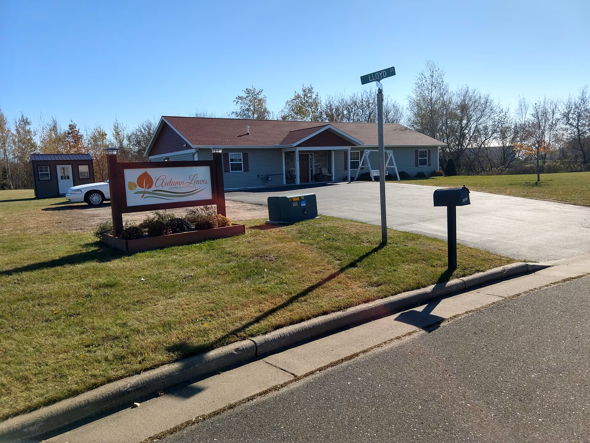 Exterior view of Autumn Leaves Assisted Living facility showing a single-story building with a red roof and gray siding. There is a sign with the facility's name in front on a grassy area, a mailbox, a street sign labeled 'Lloyd St', and a driveway leading to the building. Trees and a clear blue sky are visible in the background.