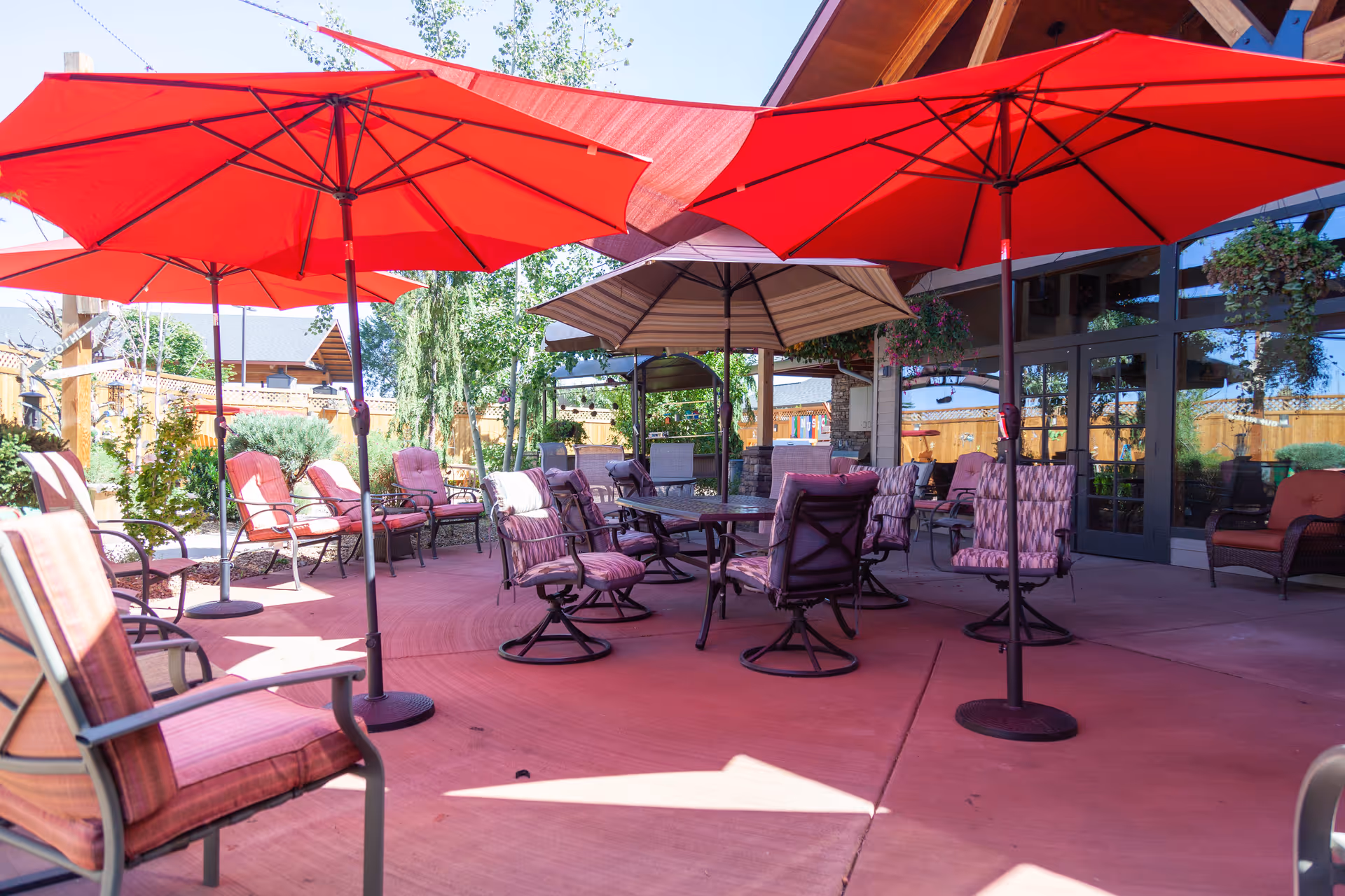 Outdoor patio area with several cushioned chairs and tables under large red and beige umbrellas. The patio is surrounded by a wooden fence and greenery, with a building featuring large glass doors and windows in the background.