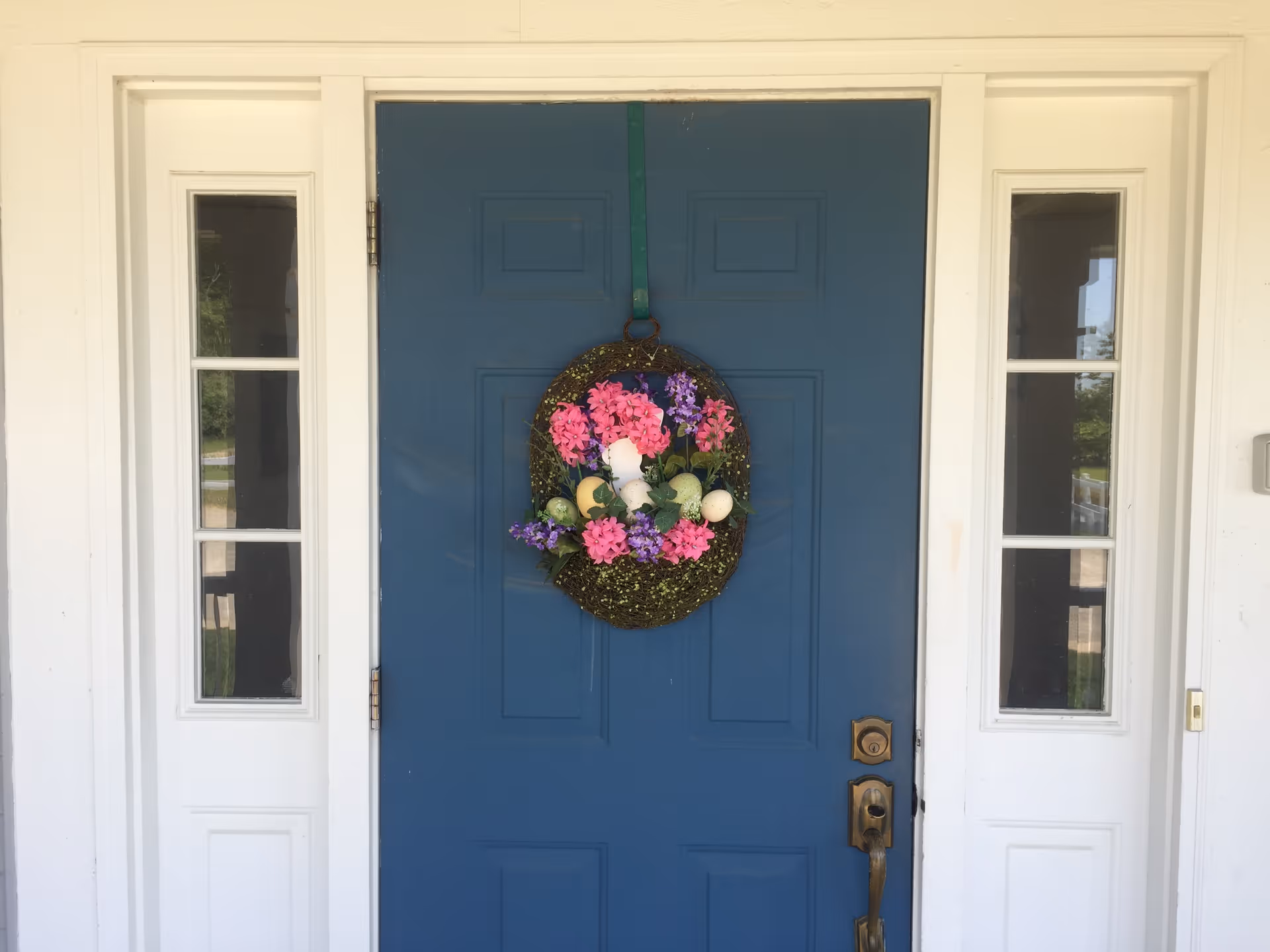 A blue front door with a decorative wreath featuring pink and purple flowers and white eggs hanging on it. The door is flanked by two narrow windows with white frames.
