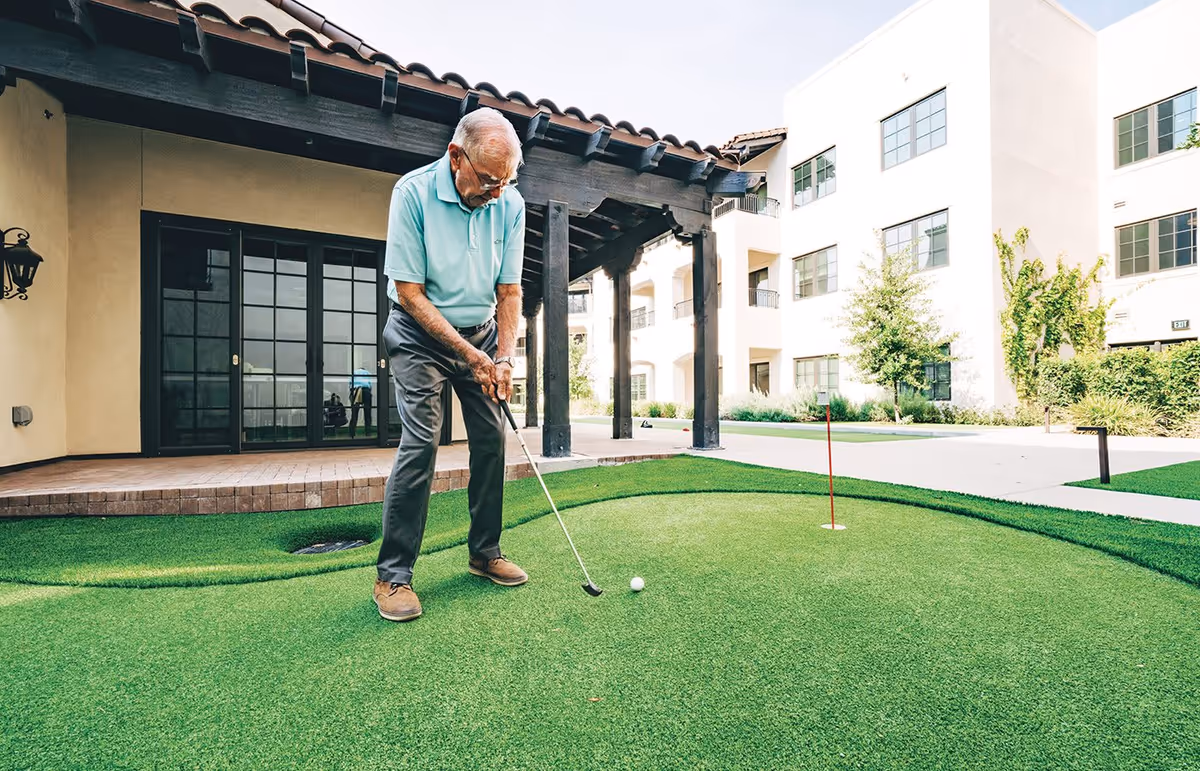 An elderly man wearing a light blue polo shirt and gray pants is playing putting golf on a small artificial green putting area outside a building with beige walls and large windows.