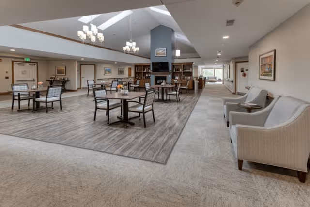 A spacious and well-lit common area in a senior living facility featuring several tables with chairs arranged on a wood-finished floor section. The room has a high ceiling with skylights and modern chandeliers. There is a fireplace with shelves and framed artwork on the walls. Comfortable armchairs are placed along the right side of the room, and a hallway extends into the distance.