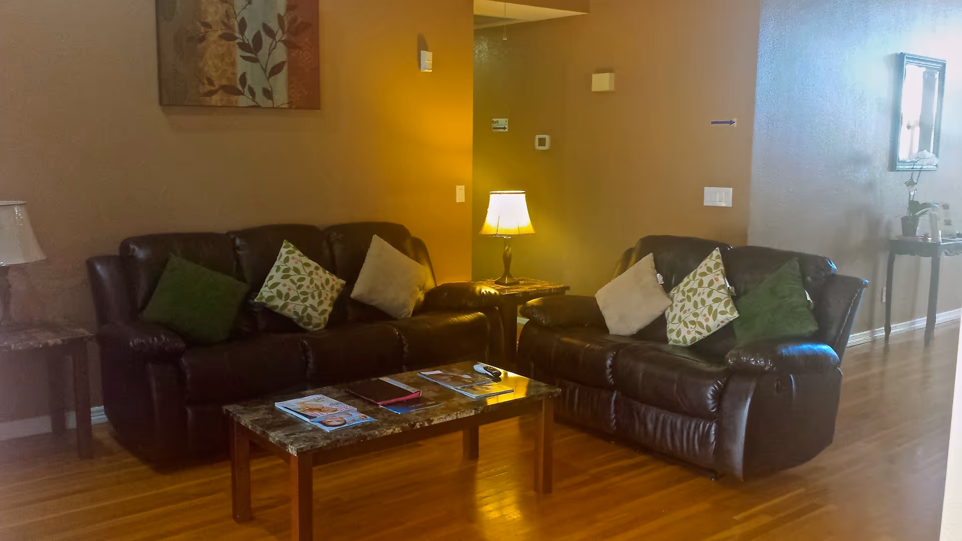 Two dark leather sofas with patterned throw pillows around a coffee table and lamp in a hardwood-floored living room.