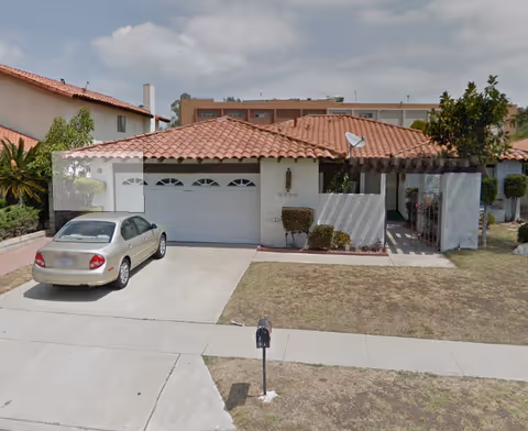 Front exterior of a single-story stucco house with a red tile roof, attached two-car garage, a car parked in the driveway, and a small front yard with a mailbox.