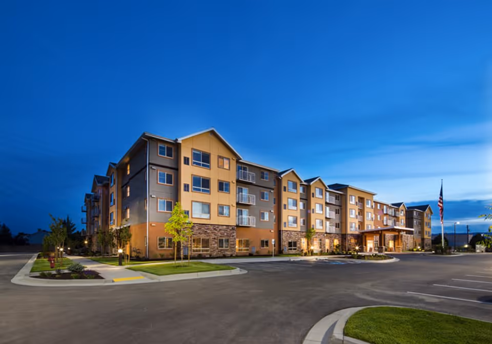 Exterior view of a modern multi-story senior living facility building at dusk with well-lit entrance, landscaped greenery, and an American flag on a flagpole in the parking area.