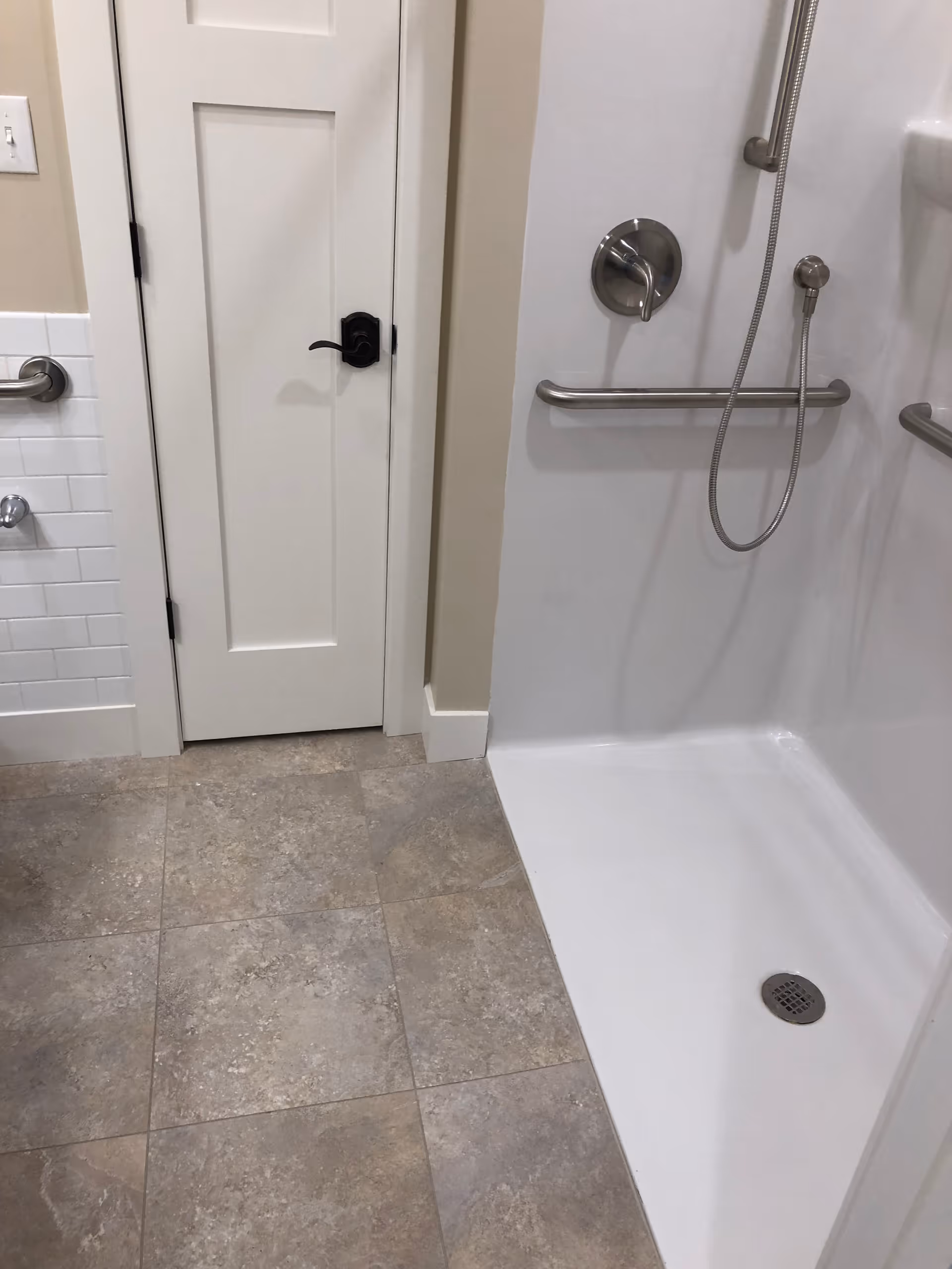A bathroom shower area with beige tiled floor, white walls, a white door with a black handle, and stainless steel grab bars. The shower has a handheld showerhead and a drain on the floor.