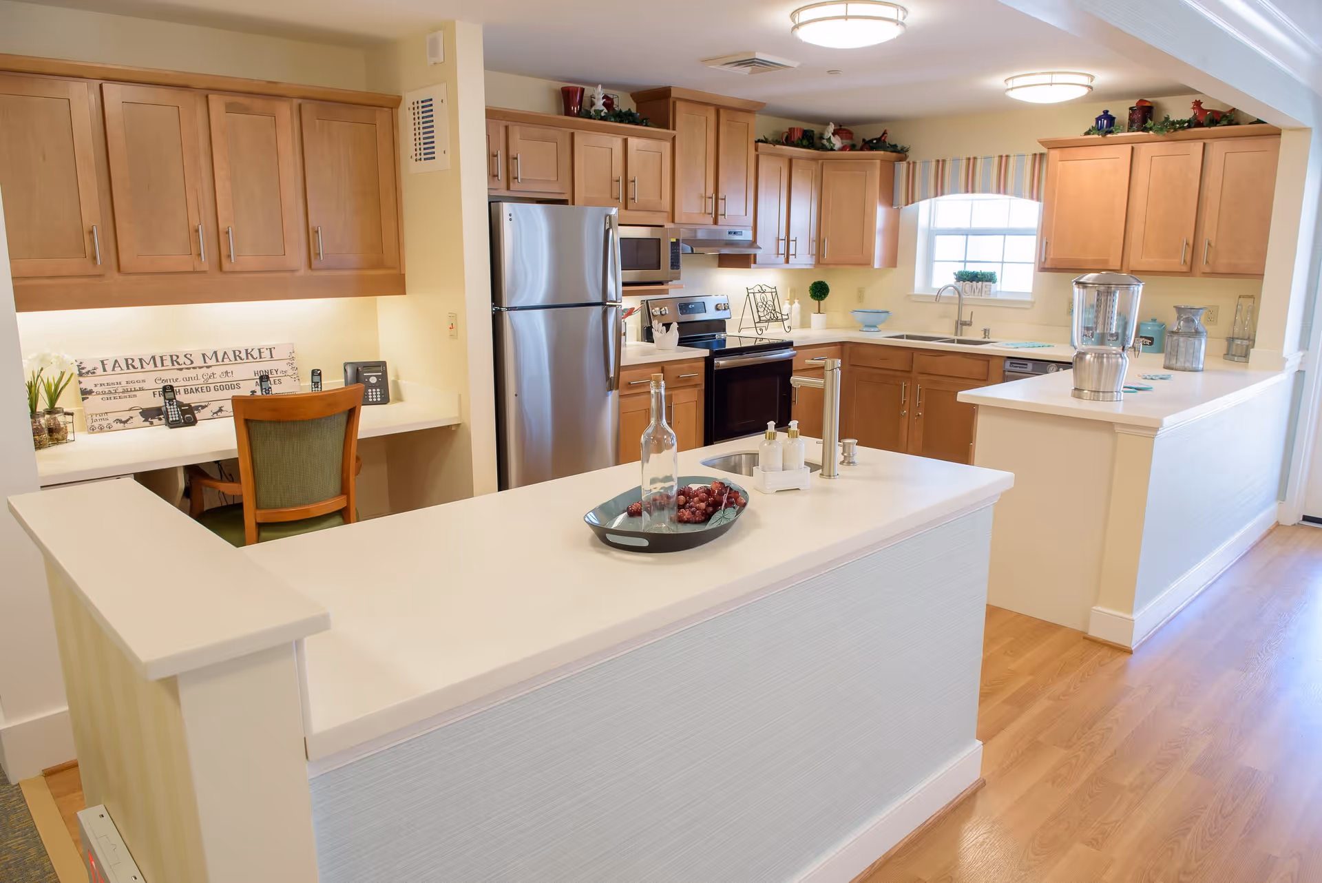 A bright and spacious kitchen area with light wood cabinets, stainless steel refrigerator, stove, and microwave. The kitchen features a large white countertop island with a decorative tray holding a bottle and grapes. There is a small desk area with a chair and a 'Farmers Market' sign on the wall. The floor is light wood, and the room is well-lit with ceiling lights and a window above the sink.