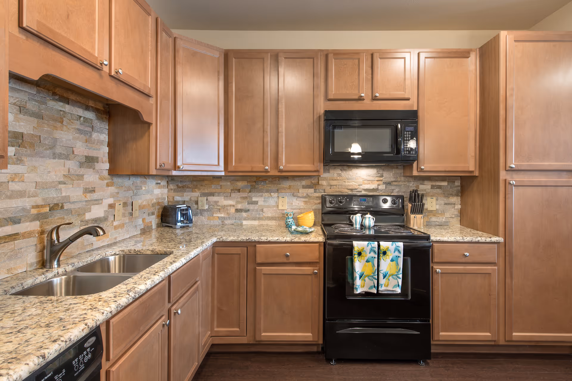 Modern kitchen with wooden cabinets, granite countertops, a double sink, black stove and microwave, and a stone tile backsplash.