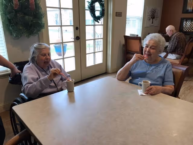 Two elderly women sitting at a table in a well-lit room, each holding a cup and stirring with a spoon. One woman is in a wheelchair wearing a light purple sweatshirt, and the other woman is wearing a light blue shirt. Behind them are glass doors with wreaths hanging on them and a man sitting at another table in the background.