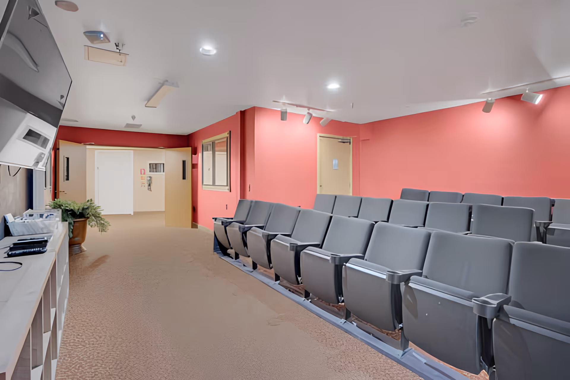 Interior view of a room with rows of gray theater-style seating facing a large screen on the left wall. The walls are painted red and beige, and the ceiling has recessed and track lighting. There is a beige carpeted floor and a door at the back of the room.