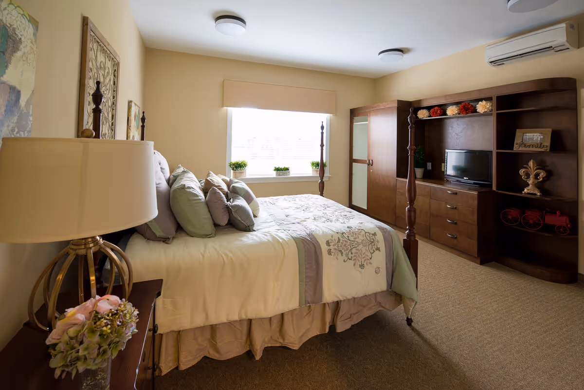 Bright, neatly arranged bedroom with a four-poster bed, bedside table and lamp, and a large wooden dresser/entertainment unit.
