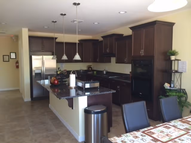 A modern kitchen with dark wood cabinets, a granite countertop island with three hanging pendant lights above, stainless steel refrigerator, built-in oven, and a dining table with chairs in the foreground.