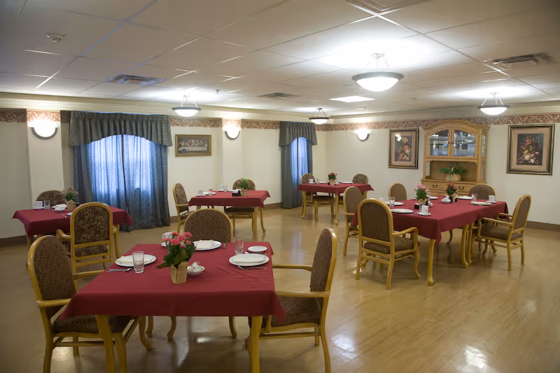 Dining room with multiple tables covered in red tablecloths, set with plates and floral centerpieces and surrounded by upholstered chairs on a hardwood floor.