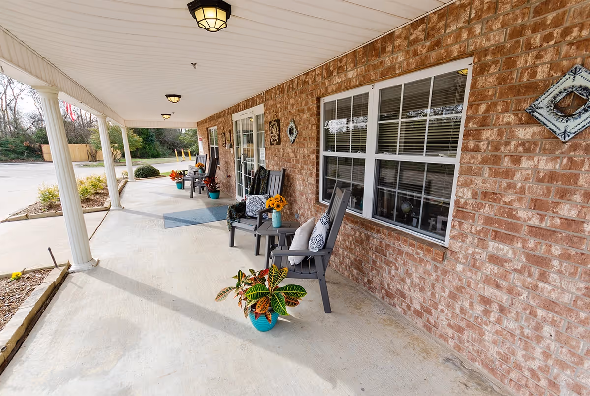 Covered outdoor porch area with brick wall, several black rocking chairs with cushions, small tables with flower vases, potted plants, and decorative wall hangings. The porch overlooks a parking area and landscaped garden beds.