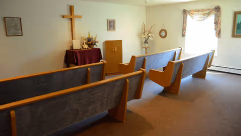 Small chapel-like room with wooden pews facing a cross, a lectern with flowers, and a window with a valance.