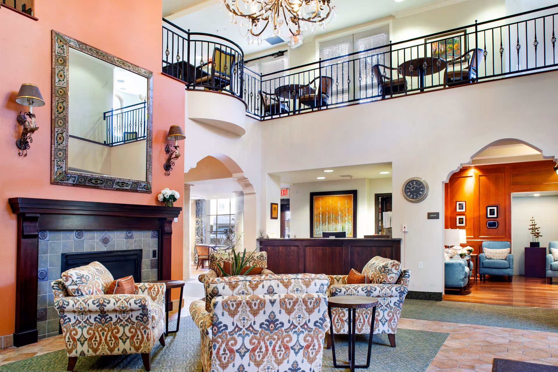 A spacious and elegant senior living facility common area with patterned armchairs arranged around a fireplace with a large decorative mirror above it. The room features a high ceiling with a chandelier, an upper balcony with seating, and an open reception desk area. Warm lighting and tasteful decor create a welcoming atmosphere.