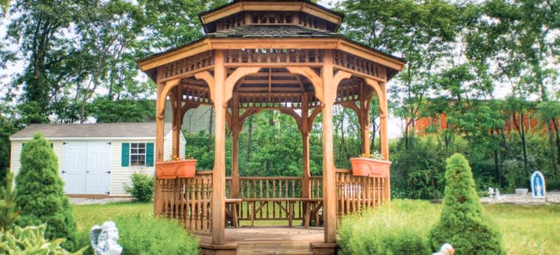 A wooden gazebo with a peaked roof and decorative railings stands in a lush green garden area surrounded by bushes, trees, and a small white shed with green shutters in the background.