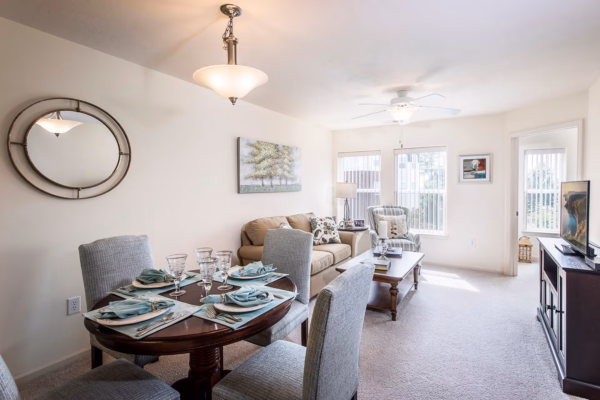 A bright and cozy living and dining area featuring a round dining table set with four place settings, a beige sofa with patterned pillows, a striped armchair, a wooden coffee table with books and decor, a TV on a dark wooden stand, and large windows letting in natural light.