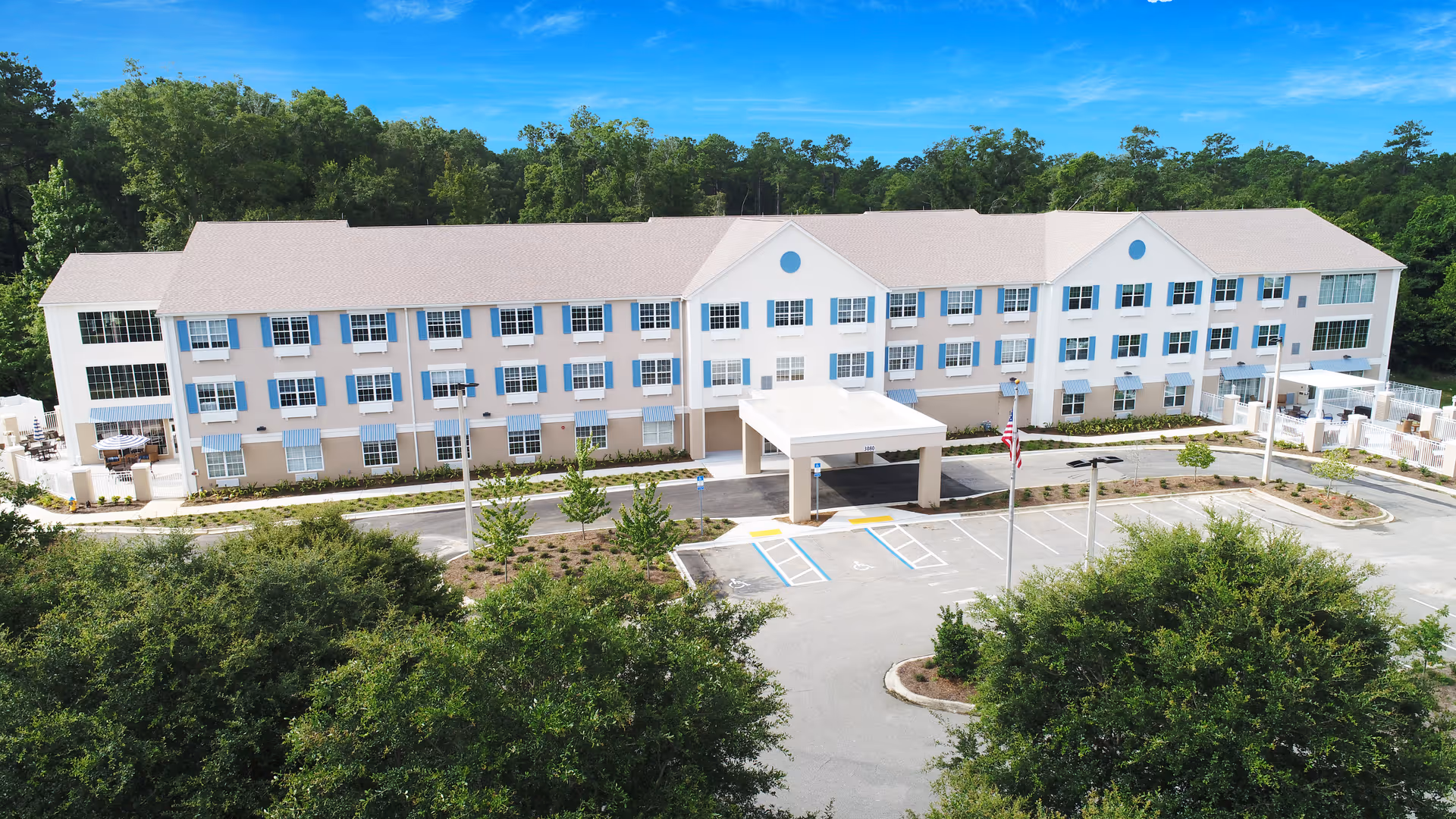 Exterior view of a large three-story senior living facility building with beige and white walls and blue window shutters, surrounded by trees and greenery, with a parking lot and entrance canopy in front under a clear blue sky.