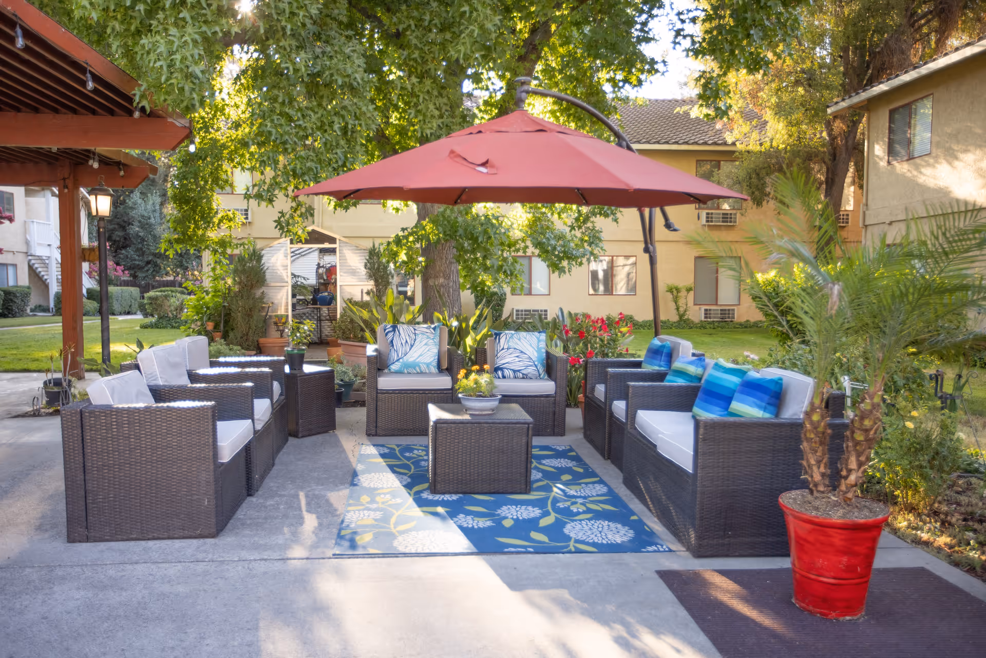 Outdoor seating area with wicker furniture including chairs and sofas with cushions, a blue patterned rug, a red umbrella providing shade, and various potted plants around. The setting is in a garden area with trees and a building in the background.