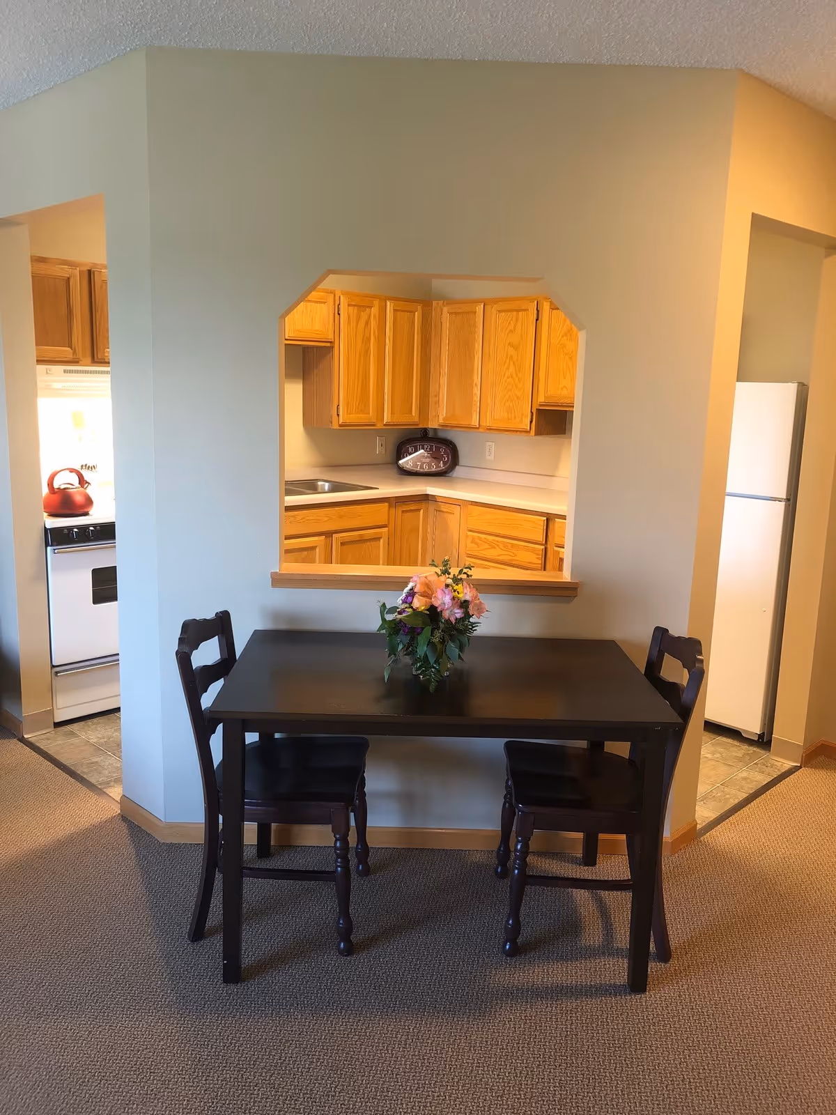 A small dining area with a dark wooden table and two matching chairs. A floral arrangement is placed in the center of the table. Behind the dining area is a pass-through window opening into a kitchen with wooden cabinets, a white countertop, a stove with a red kettle, and a white refrigerator.