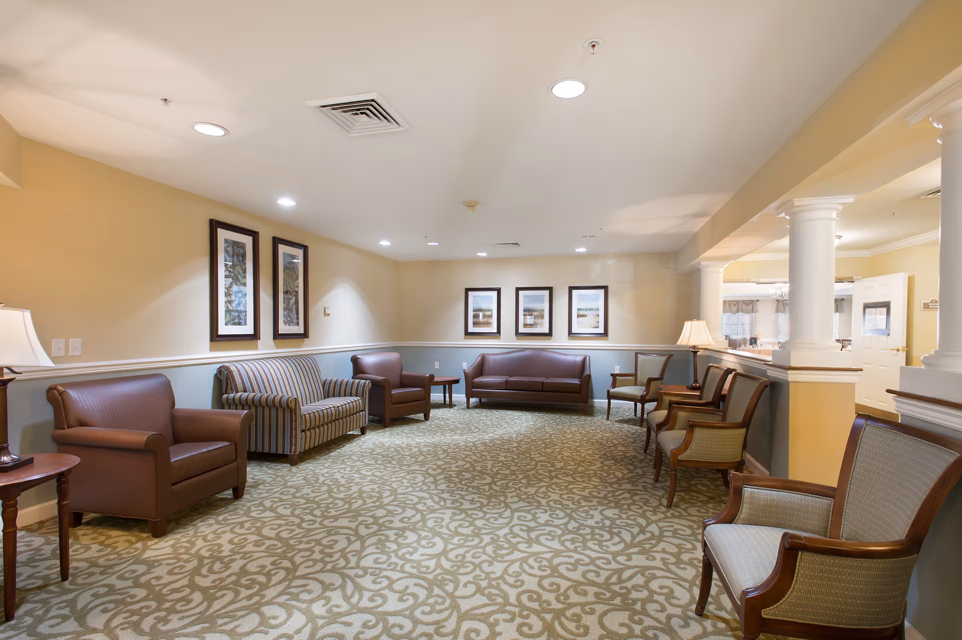 Well-lit lounge in a senior living facility with sofas and armchairs arranged along the walls, patterned carpet, and framed artwork.