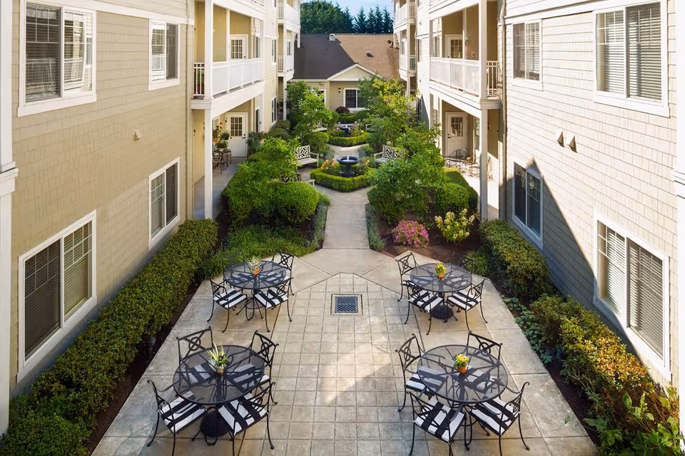 Landscaped courtyard between two apartment buildings featuring tiled patios with round metal tables and chairs, shrubs, and a central fountain.
