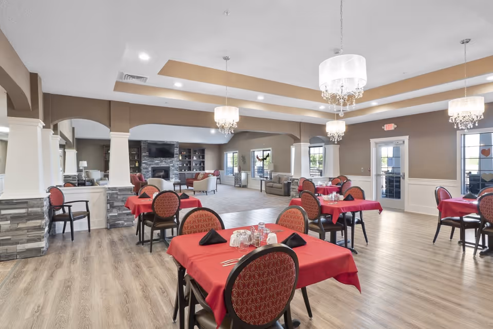 A spacious dining room with several tables covered in red tablecloths and set with black napkins, glasses, and silverware. The room features wooden flooring, elegant chandeliers hanging from the ceiling, and a seating area with armchairs and a fireplace in the background. Large windows and glass doors allow natural light to enter the space.