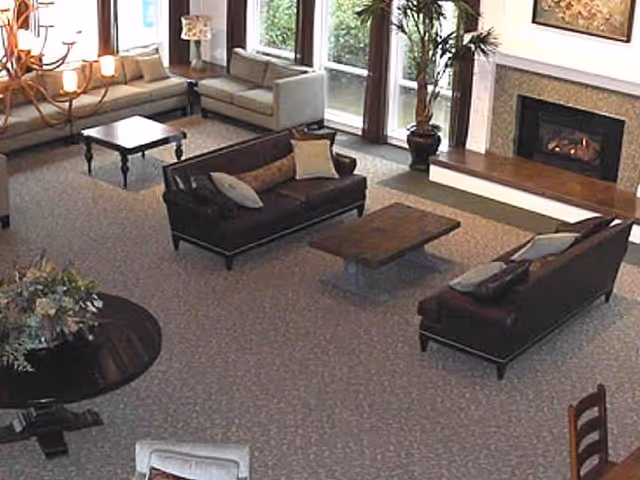 A spacious living room area with multiple sofas and armchairs arranged around two wooden coffee tables. There is a fireplace on the far wall with a painting above it, large windows letting in natural light, a potted plant near the fireplace, and a round table with a floral arrangement in the foreground.