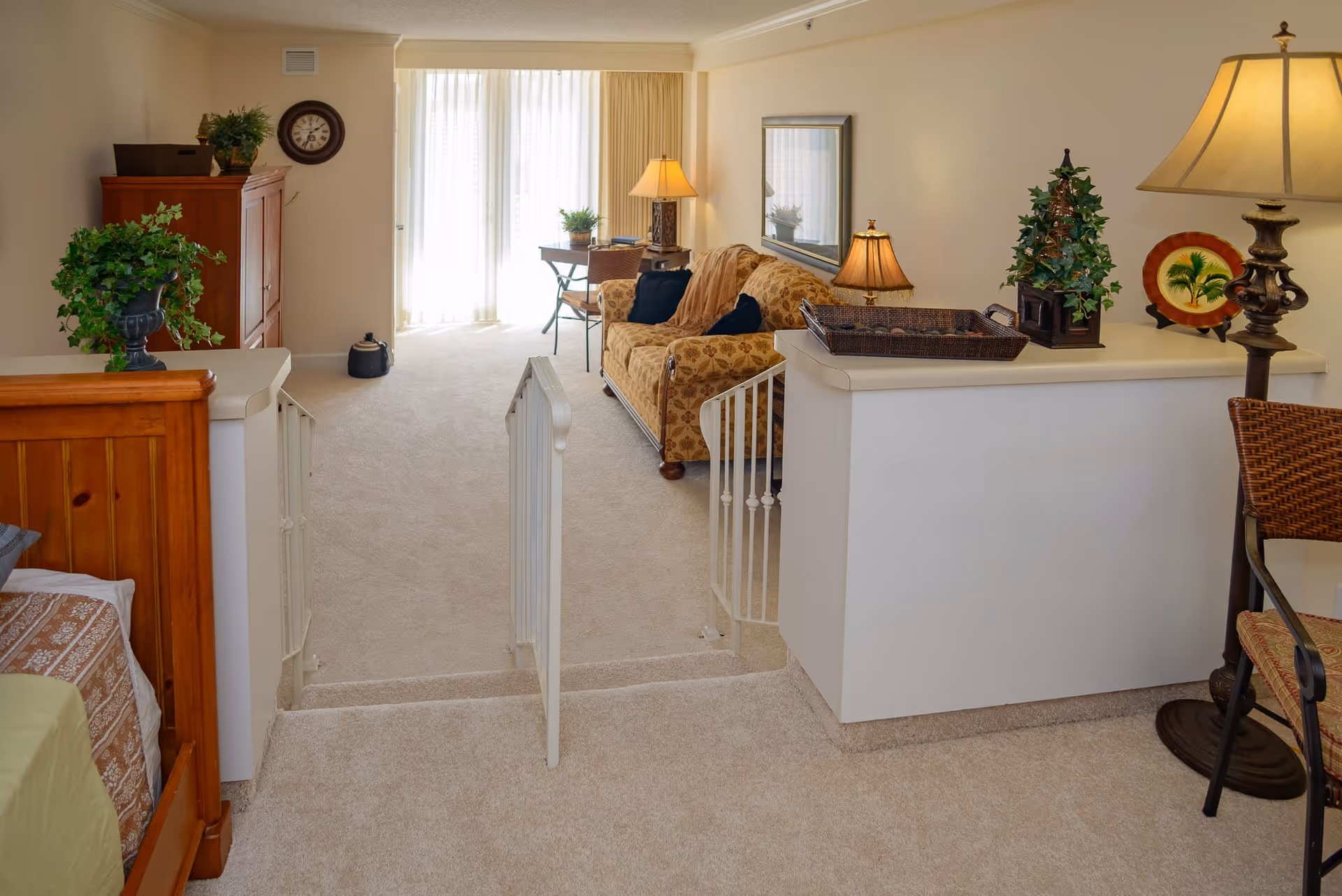 Interior view of a senior living facility room at Garden Plaza at Inverrary featuring a cozy living area with a patterned sofa, two table lamps, a small desk with a chair near a window with sheer curtains, and a wooden bed partially visible on the left. The room has beige carpeting and light-colored walls, with decorative plants and framed artwork adding warmth to the space.