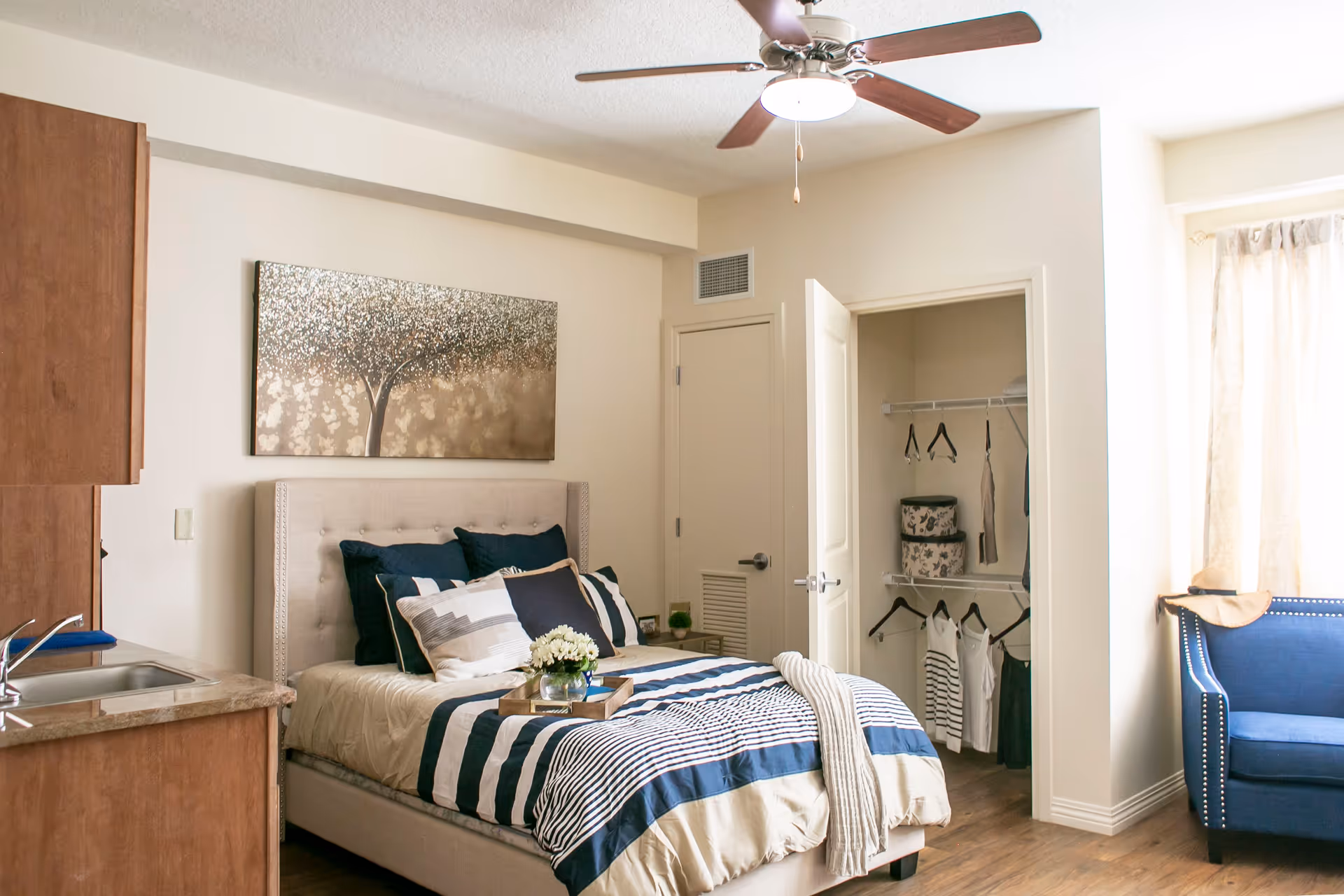 A cozy bedroom in a senior living facility featuring a bed with striped bedding and multiple pillows, a small wooden side table with a flower vase, an open closet with hanging clothes and storage boxes, a blue armchair near a window with light curtains, a ceiling fan with wooden blades, and a kitchenette area with a sink and wooden cabinets.