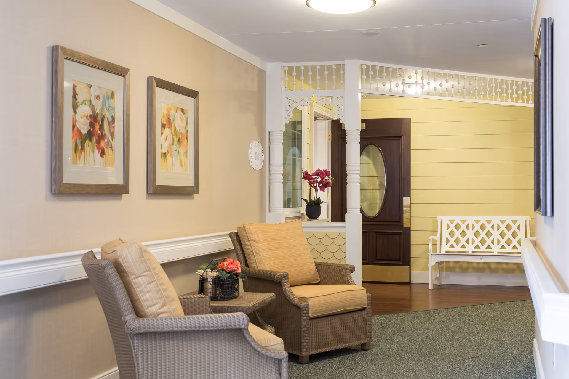A cozy seating area in a senior living facility hallway with two cushioned wicker chairs and a small table with a flower arrangement. The walls are decorated with framed floral paintings, and there is a white bench near a wooden door with an oval glass window. The area has warm lighting and a welcoming atmosphere.