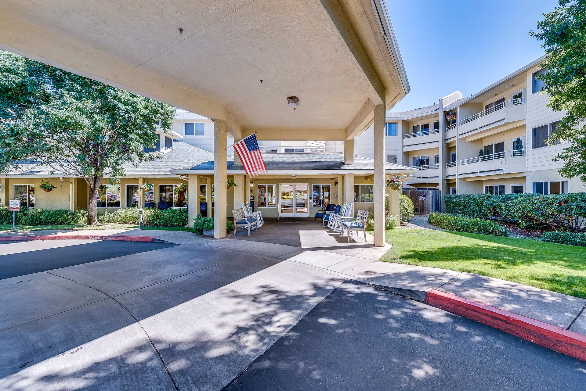 Entrance of Solstice Senior Living At Auburn showing a covered drop-off area with benches and rocking chairs, an American flag hanging from the ceiling, surrounded by a three-story building with balconies, trees, and well-maintained landscaping under a clear blue sky.