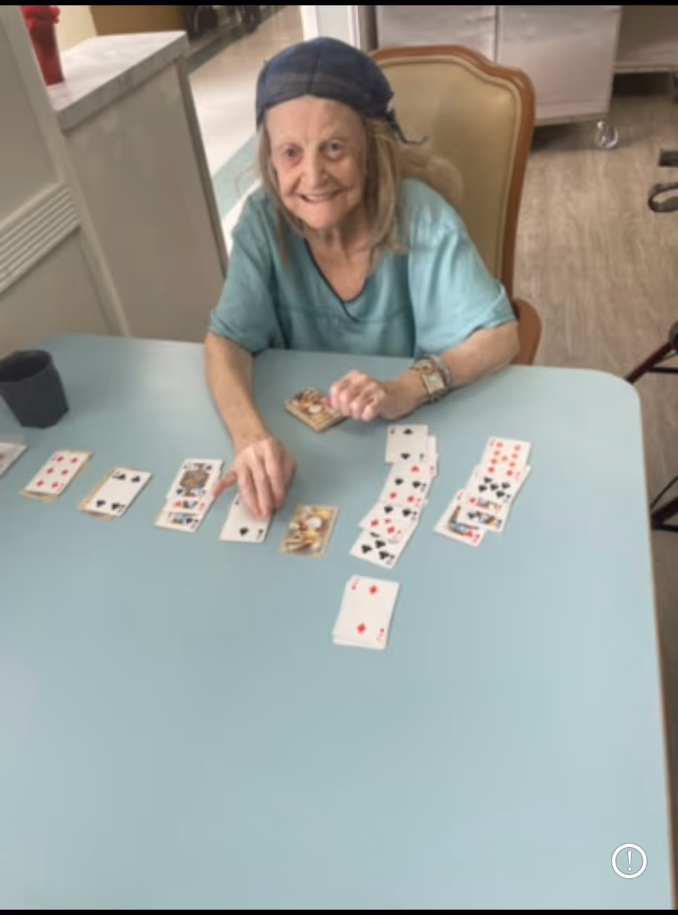 An elderly woman wearing a blue headscarf and teal shirt is sitting at a light blue table playing a card game. Several playing cards are spread out on the table in front of her. The setting appears to be indoors with light-colored flooring and furniture visible in the background.